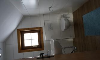 A small, modern bathroom with a white wall design and wooden floor. It contains a sink, a toilet, and a window with a wooden frame.