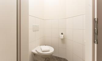 A small, modern bathroom with white tiles and a toilet. A towel is on the shelf, and a toilet paper holder is available.