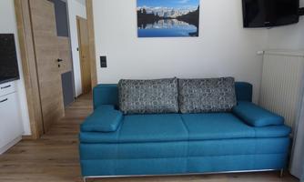 A modern living room with a turquoise sofa and a television mounted on the wall. In the background, there is a picture of a mountain landscape over a tranquil lake.