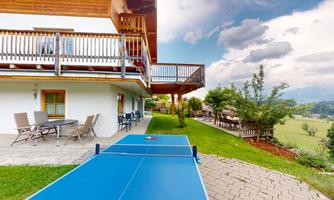 A terrace with a table tennis table and comfortable seating. In the background, there is a green lawn and a cloudy sky.