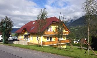 A yellow house with a red roof stands at the roadside. In the background, green mountains and trees are visible.