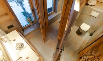 A modern bathroom with wood details and large windows. There are two sinks and a toilet, surrounded by stylish floor tiles.