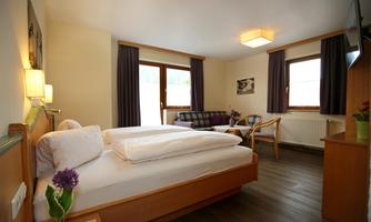 A cozy room with a double bed, two windows, and a seating area. The decor is modern and inviting, featuring wooden furniture and soft colors.