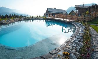 A clear pool surrounded by green grass and flowers. In the background, rustic buildings and mountains can be seen.