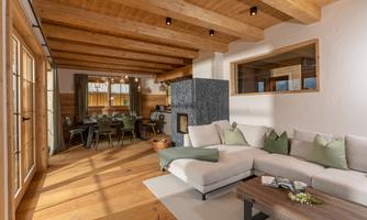 A bright and cozy apartment with wooden panels and large windows. The open living area includes a corner sofa, a coffee table, and a dining area in the background.