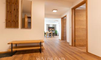 An inviting hallway with a wooden shoe rack and mirror. In the background, a bright living area with plants is visible.