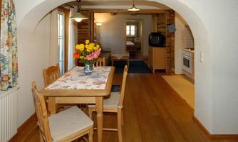 A cozy dining area with a wooden table and chairs. A vase with colorful flowers is on the table, in the background you can see the kitchen and a living area.