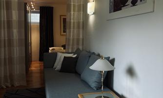 A modern living room with a blue sofa and a small table. The walls are light, and there are artworks and curtains that create a cozy atmosphere.