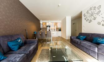 A modern furnished living room with two sofas and a glass table. The walls are decorated in warm colors, and the kitchen is open and well-lit.