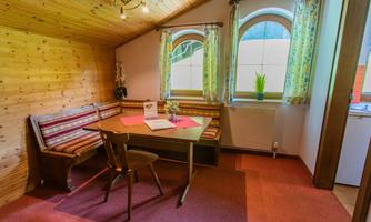 A cozy room with wooden walls and colorful curtains. A table with chairs is in the center, and large windows let in plenty of light.