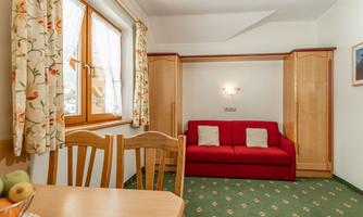 A cozy living space with a red sofa and a wooden table. The windows are adorned with curtains and the floor is covered with green carpet.