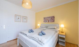 A cozy bedroom with a large bed and two nightstands. The walls are painted yellow and there are decorative pictures as well as warm light sources.