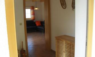A bright entrance area with a wooden cabinet and ceramic tiles. In the background, a living room with a sofa and a window can be seen.