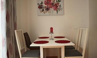 A bright dining area with a white table and four chairs. In the center, there is a red candle, and on the wall hangs a floral picture.