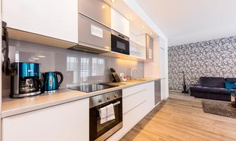 A modern kitchen with elegant gray furniture and integrated appliances. In the background, there is a cozy living room with a floral patterned wall.