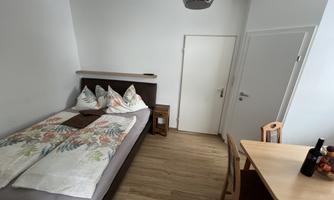A bright room with a single bed and a small table. The walls are white and the floor is wooden.