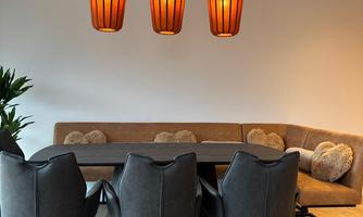 A modern dining room with an oval table and stylish black chairs. Three orange lamps hang above the table, and a cozy seating area is visible in the background.