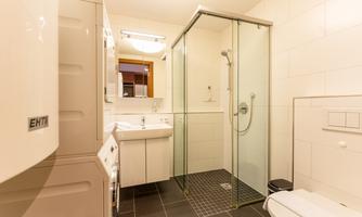 A modern bathroom with a shower, a sink, and a washing machine. The walls are tiled in light colors, and the floor is in a dark color.