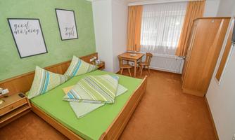 A cozy bedroom with a double bed and colorful pillows. There is a small table and a chair, as well as large windows with curtains.
