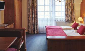 A cozy hotel room with wooden furniture and a double bed. Bright light streams in from a large window and the curtains bring color into the room.