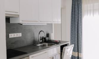 A modern kitchen with white cabinets and a gray workspace. It includes a stove, a refrigerator, and large windows with curtains.