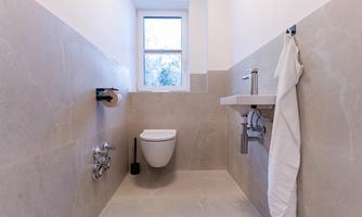 A modern bathroom with bright natural stone, a toilet, and a small sink. A window provides daylight and a view outside.