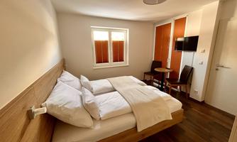 A bright bedroom with a double bed and a small table. The room has wooden floors and large windows with blinds.