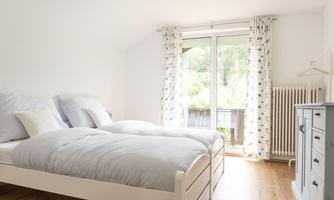 A bright bedroom with two single beds and a wooden floor. Large windows offer a view of the greenery.