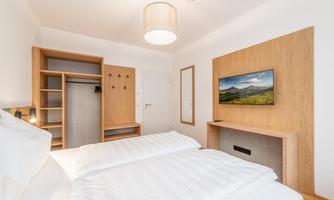 A modern bedroom with two single beds and wooden furniture. A small television hangs on the wall, and the room feels bright and inviting.