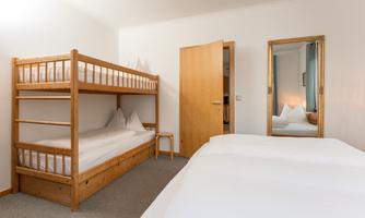 A simple bedroom with a bunk bed and a double bed. The walls are light and there is a mirror on the wall.