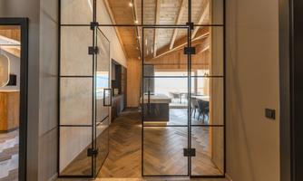 A modern entrance area with glass doors and wooden paneling. In the background, a stylish living and dining room with a view of nature can be seen.