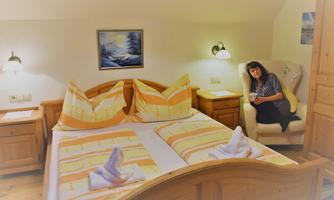 A cozy room with a large bed and wooden furniture. A person is sitting in an armchair and smiling.