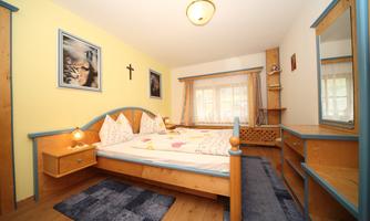 A cozy bedroom with a double bed, wooden furniture, and colorful bedding. The room has large windows and a warm atmosphere.