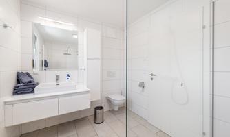 A modern bathroom with white tiles and a clear, bright design. It features a shower, a sink with a mirror, and a toilet.