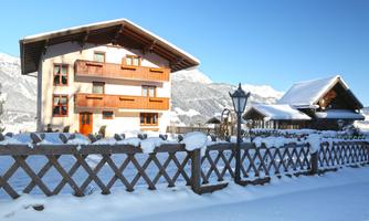 A beautiful, snow-covered house with traditional architectural elements. The garden is surrounded by a wooden fence, and the mountains are visible in the background.