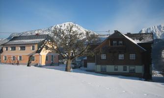 A quiet winter village with snow-covered houses and a large tree. In the background, mountains are visible under a clear blue sky.