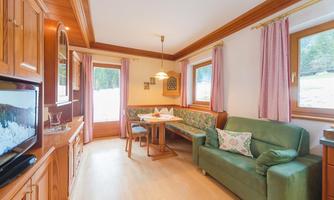 A cozy living space with wooden paneling and a green sofa. A table with chairs is located in the middle, and large windows offer a view outside.