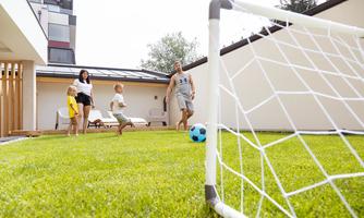 A cheerful game of children in the garden with a soccer goal. In the background, there are some lounge furniture and a modern house visible.