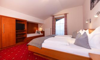A cozy bedroom with two beds and modern wooden decor. The windows bring plenty of daylight into the room, which is carpeted with a red rug.