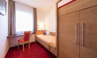 A simple room with a single bed and a desk. The room has light walls and a wooden cupboard.