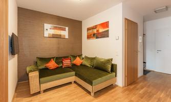 A cozy living room with a green couch and colorful cushions. Modern artworks hang on the wall and the floor is made of light wood.