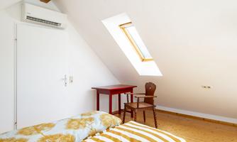 A cozy room with wooden floors and a skylight. There is a bed with striped blankets, a small table, and a chair.