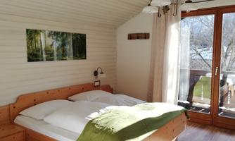 A cozy bedroom with a double bed and bright wood interior. Large windows offer a view of a balcony and plenty of daylight.