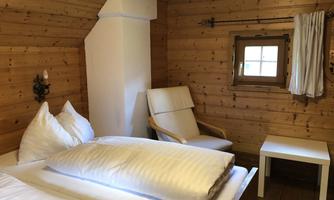 A cozy room with wood paneling and a large window. There is a bed with white pillows and a corner with an armchair and a small table.