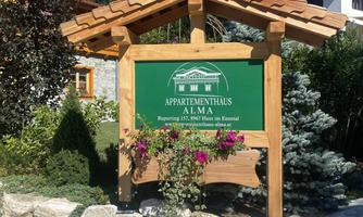 A wooden sign with the inscription "Apartment House Alma" in a picturesque setting. Surrounded by greenery and rocks, the place exudes an inviting atmosphere.