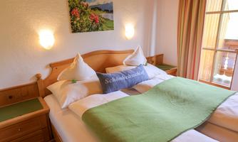 A cozy bedroom with a double bed, white bedcovers, and a green throw. A landscape painting hangs on the wall, and there are light curtains that let in some daylight.
