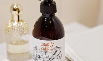 An elegant dispenser with shampoo in a brown bottle stands on a bathroom shelf. In the background, a perfume bottle and a towel can be seen.