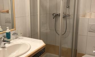 A bathroom with a shower cabin and a sink. The walls are tiled white and the floor has a beige color.