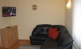 A cozy living space with a black sofa and a red cushion. A television is hanging on the wall and there is a wooden cabinet.