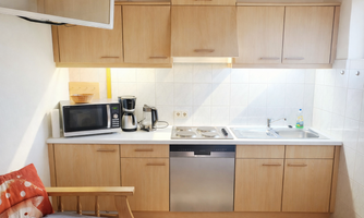 A modern kitchen with light wooden cabinets and a countertop. The equipment includes a refrigerator, a microwave, a dishwasher, and a coffee machine.